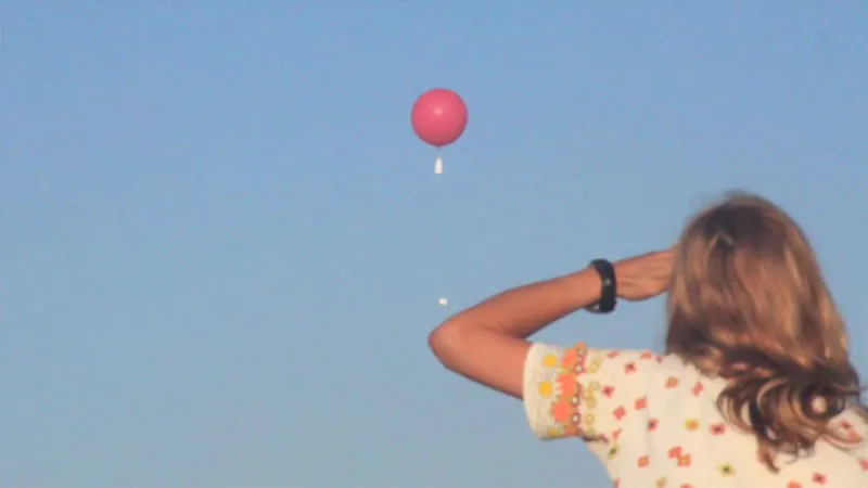 A photo of a woman looking up at a giant red helium balloon floating in a cloudless sky.